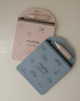 The mushie Reusable Water Color Pad includes two spiral-bound pads (pink and blue) with line drawings and refillable paintbrushes on top. “mushie” is printed on each eco-friendly cover, making sustainable art fun for kids.
