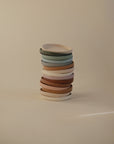 A vertical stack of Mushie Classic Silicone Suction Plates in minimalist pastel shades—white, beige, brown, and muted green—stands against a plain light beige background. The non-toxic toddler plates feature a clean, modern design.