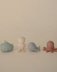 Four pastel silicone bath toys—a blue submarine, cream diver, gray whale, and dusty pink octopus—from the mushie Sealife Mold Free Bath Play Set are lined up on a plain surface against a beige background.