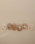 The FRIGG Natural Rubber 6-Pack by FRIGG features six neutral-toned (cream, taupe, brown) pacifiers with round handles and subtle edge variations, all made in Denmark and shown against a plain beige background.