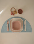 The mushie Dinnerware Cutlery Set features a pink plastic fork, knife, bowl, and cup on a pastel blue semicircular placemat with a beige teether toy placed above the utensils—all arranged neatly on a light surface.