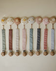 Mushie Linen Pacifier Clip 2-Pack, pastel colors, wooden clips on beige background.