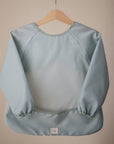 The mushie Long Sleeve Bib in light blue, featuring elastic cuffs and a water-resistant, wipe-clean surface, hangs on a wooden hanger against a beige background with a small white label at the bottom center.