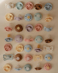 A grid of FRIGG Daisy Silicone Pacifier 2-Pack | Limited Edition by FRIGG, featuring colorful pastel designs in pink, blue, yellow, and brown for baby comfort, arranged neatly on a light beige background.