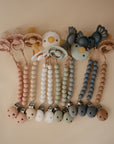 Five mushie Hera Silicone Pacifier Clips in soft pink, cream, green, gray, and brown are arranged in a row. Each features food-grade silicone and minimalist flower or butterfly designs in pastel colors.