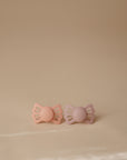 Two FRIGG Lucky Symmetrical Silicone Pacifiers (6-18 Months), in peach and lavender, are placed side by side on a smooth beige surface. The butterfly-shaped design and medical grade silicone nipple ensure comfort for your baby.