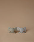 The FRIGG Butterfly Anatomical Silicone Pacifier 2-Pack (0-6 Months) features two butterfly-shaped pacifiers in sage green and light grey, displayed side by side on a plain, light brown background.