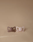 The FRIGG Butterfly Anatomical Silicone Pacifier 2-Pack (6-18 Months) features mauve and light pink pacifiers, placed side by side on a beige surface with a matching background for gentle baby comfort.