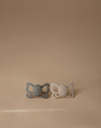 The FRIGG Butterfly Anatomical Silicone Pacifier 2-Pack (6-18 Months) features gray and beige pacifiers with a butterfly-shaped shield, displayed side by side on a light beige surface against a plain beige background for baby comfort.