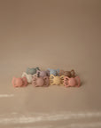 Nine pastel FRIGG Lucky Symmetrical Silicone Pacifiers (6-18 Months) are grouped on a beige surface with soft shadows, showcasing the high-quality design by FRIGG—ideal for supporting your baby's oral development. Each pack contains 2 pacifiers.
