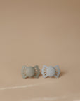 Two FRIGG Lucky Symmetrical Silicone Pacifiers (0-6 Months), one green and one light blue, with medical grade silicone nipples, are placed side by side on a smooth beige surface with a soft gradient background.