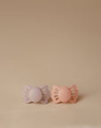 The FRIGG Lucky Symmetrical Silicone Pacifier 2-Pack (0-6 Months) features two butterfly-shaped pacifiers in light purple and peach, shown side by side on a beige background, designed for baby comfort and safety.