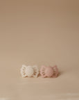 Two FRIGG Lucky Symmetrical Silicone Pacifiers (0-6 Months) in white and light pink, both with medical-grade silicone nipples and butterfly-shaped shields, are side by side on a beige surface with a plain beige background.