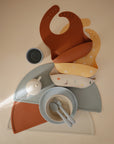 A flat lay of mushie baby feeding essentials—food-grade silicone bibs, bowl, cup, plate, spoon, fork, and two Silicone Placemats—arranged neatly in muted earth tones and soft pastels on a beige surface.