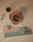 A neatly arranged set of mushie Square Dinnerware Plates, BPA-free and pastel-colored, displayed with matching bowls, cups, baby plates, spoons, bottle, and placemats on a light surface.