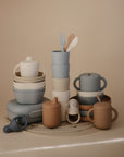 Mushie Dinnerware Cups and other pastel silicone baby feeding items—non-toxic cups, bowls, plates, fork, spoon, and pacifier—are neatly arranged on a beige surface as perfect alternatives to plastic dinnerware.