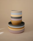 A stack of mushie Round Dinnerware Bowls and plates in yellow, pink, gray, and light blue—BPA-free pastel-colored plastic—arranged neatly against a plain beige background.