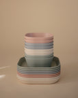 A neatly stacked set of mushie Square Dinnerware Plates in pastel pink, blue, green, and white, made from BPA-free plastic and arranged in alternating layers against a beige background.