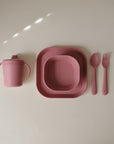 The mushie Dinnerware Fork and Spoon Set is a pink, non-toxic children’s dining set with a dishwasher-safe sippy cup with handles, two square plates, plus a plastic fork and spoon—all arranged neatly on a light surface.