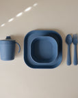 The mushie Square Dinnerware Bowls set features blue silicone BPA-free kids’ tableware—a sippy cup with handles, square bowl, plate, fork, and spoon—neatly displayed on a beige surface.