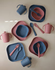 Four mushie Square Dinnerware Plates sets in BPA-free plastic, each with a plate, bowl, fork, spoon, and sippy cup in muted pink and blue tones, are neatly arranged on a neutral background.