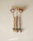 Three pastel baby pacifiers (cream, beige, mauve) are attached to mushie Silicone Pacifier Clip | Hera, featuring a minimalist design and arranged vertically on a light, neutral background.