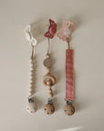 Three mushie Silicone Pacifier Clip | Hera designs are shown: two with minimalist wooden beads, and one with a pink silicone ribbon. Each has butterfly or flower shapes and secure food-grade silicone holders.