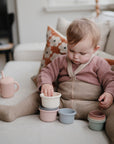 A toddler in a pink sweater, taupe tights, and brown shorts stacks mushie Stacking Cups Toy on the couch. A pink sippy cup sits nearby, with a floral pillow in the background.