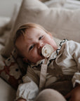 A baby with light hair lies on a couch, smiling with a FRIGG Rope Silicone Pacifier in their mouth. They wear a white ruffled shirt and beige overalls, resting against a floral pillow. The pacifier is from the FRIGG Rope Silicone Pacifier 2-Pack.
