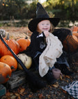 A young child in a witch costume sits among pumpkins, smiling and holding a mushie Organic Cotton Muslin Cloth from the 3-Pack set. Surrounded by festive Halloween props, it's perfect for your nursery needs!.