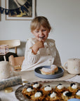 Happy child enjoys a donut on a Mushie Classic Silicone Suction Plate, snacks and drinks close by.