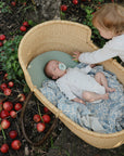 A baby in a basket with a mushie Organic Cotton Muslin Swaddle; apples scattered; toddler reaching.
