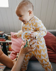 Baby in heart PJs holds mushie Teething Gift Set while being lifted by an adult on the couch.