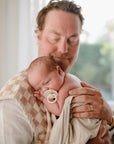 A man gently holds a sleeping newborn on his shoulder, resting on mushie’s Organic Cotton Muslin Burp Cloth 2-Pack. The baby uses a pacifier and is wrapped in a light blanket as soft natural light streams through the window.