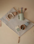 The mushie Silicone Toddler Starter Spoons, in soothing pastel colors, are arranged neatly on a beige ribbed cloth—paired with bowls, cups, a toy phone, and pacifiers—creating an inviting setup for independent feeding.