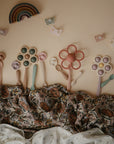 Mushie Silicone Toddler Starter Spoons are artfully arranged with colorful flower-shaped toys on a floral-patterned blanket, while playful butterflies and a rainbow complete the charming, neutral-toned setting.