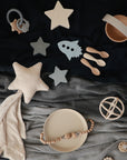 A flat lay of neutral baby items on fabric, including star-shaped toys, a rocket, mushie Silicone Toddler Starter Spoons for independent feeding, a soft star pillow with cloth, a plate, teether, rattle, and bowl.