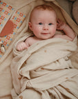 A light-skinned baby with red hair, wrapped in a beige rainbow-patterned blanket, smiles up while lying next to a colorful mushie Water Resistant Wet Bag and baby accessories.