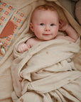 A light-skinned baby with red hair, wrapped in a beige rainbow-patterned blanket, smiles up while lying next to a colorful mushie Water Resistant Wet Bag and baby accessories.