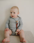Baby holds a mushie Linen Pacifier Clip 2-Pack, wearing gray onesie, looking to the side.