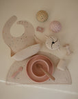 The mushie Silicone Placemat set in pastel includes a food-grade speckled bib, divided plate, bowl, spoon, cup, toy food items—all arranged neatly on a matching silicone placemat.