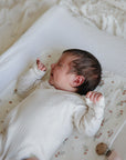 Newborn in white onesie on mushie Extra Soft Muslin Changing Pad Cover, hand raised near face.