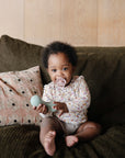 A baby with a FRIGG Andersen Fairytale Silicone Pacifier sits on a dark green couch, holding a light blue rattle. A patterned pillow is beside the baby and a light wooden wall is behind. The pacifier by FRIGG is made in Denmark from safe silicone.
