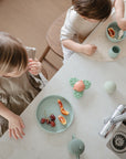 Two small children sit at a kitchen counter with snacks like grapes and orange slices served on pastel-colored Mushie Classic Silicone Suction Plates. They reach for food as small toys are scattered nearby.