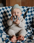 A smiling baby in a beige knit outfit and green beanie sits on a blue and white checkered blanket, holding the mushie Space Teething Ring, made from food-grade silicone to support sensory development.