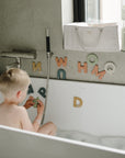 A mushie Diaper Caddy sits by the window as a young child plays with foam letters in the tub.
