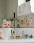 Smiling child enjoys bath with bubbles, colorful letters, and a Mushie Bath Rinse Cup nearby.