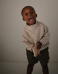 A young child smiles while standing against a plain background, wearing a striped pullover and dark shorts, holding the mushie Phone Press Toy—designed to encourage fine motor skills.