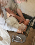 Two adults gently bathe a baby in a kitchen sink. The baby is wrapped in a mushie Organic Cotton Muslin Swaddle Blanket and looks up at one adult while the other pours water. A pacifier clip and washcloth rest on the counter.