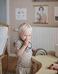 A blond-haired child stands by a table, smiling while holding toys. On the table are a wooden rainbow toy, shape sorter, and the mushie Space Teething Ring for sensory development. Neutral decor and a wicker chair complete the room’s look.